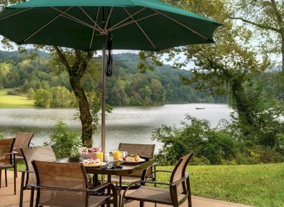 A scenic outdoor dining setup with tables, chairs, and a green umbrella overlooking a serene lake surrounded by lush greenery.