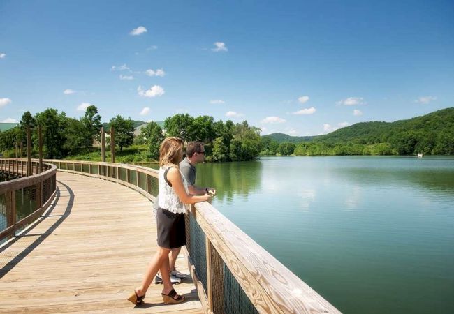 A couple stands on a wooden boardwalk overlooking a serene lake surrounded by lush green hills under a clear blue sky.