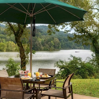 Outdoor dining setup under a green umbrella by a lake, surrounded by trees, with food and drinks on the table.