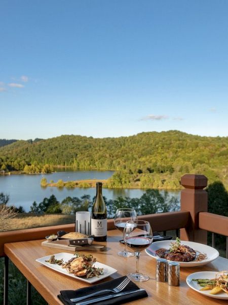 A wooden table with gourmet meals and wine overlooks a scenic lake and forested hills under a clear blue sky.