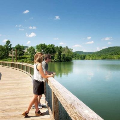 A couple stands on a wooden boardwalk overlooking a calm lake, surrounded by lush greenery and hills under a bright blue sky with scattered clouds.