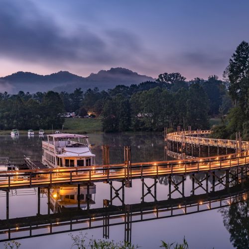 A serene landscape featuring a luminous footbridge over a calm river with a boat, surrounded by trees and mountains in the background.