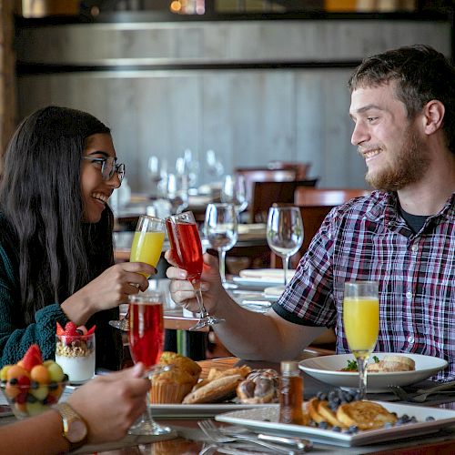 People sitting at a table in a restaurant, enjoying food and drinks, while smiling and toasting with glasses.