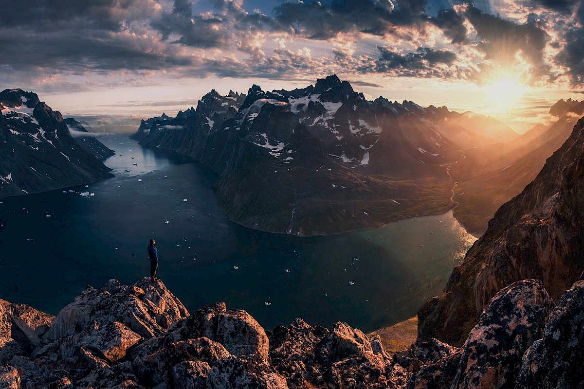 A person stands on a rocky ledge overlooking a breathtaking fjord with snowy mountains and a dramatic sunset in the background.