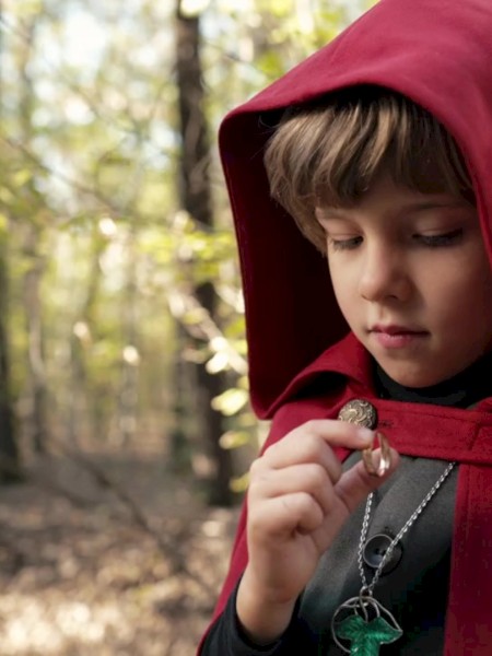 A child in a red hood stands in the forest, examining a necklace.