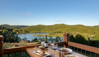 A table with dishes and wine overlooks a scenic view of a lake and forested hills under a clear sky.