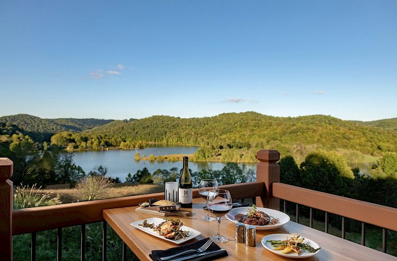 A table with dishes and wine overlooks a scenic view of a lake and forested hills under a clear sky.