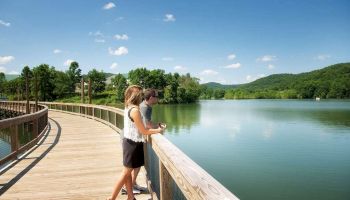 A couple stands on a wooden boardwalk overlooking a calm lake, surrounded by lush green hills under a bright blue sky with a few clouds.