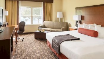 A hotel room with a bed, sofa, desk, and TV; features warm decor, natural light from the window, and modern furnishings.