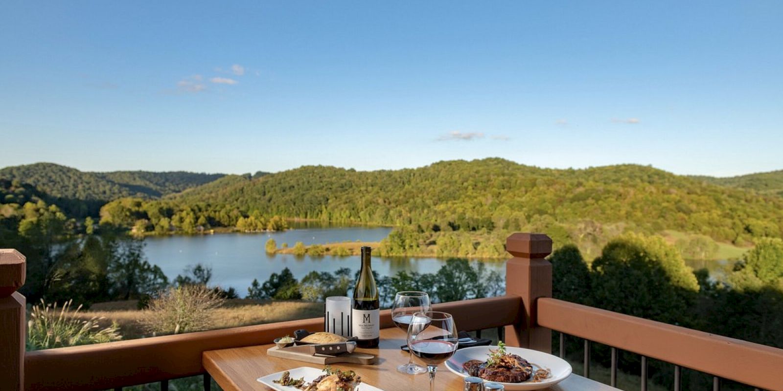 A scenic view of a lake and hills with a table set for a meal, featuring wine and dishes, on a balcony.