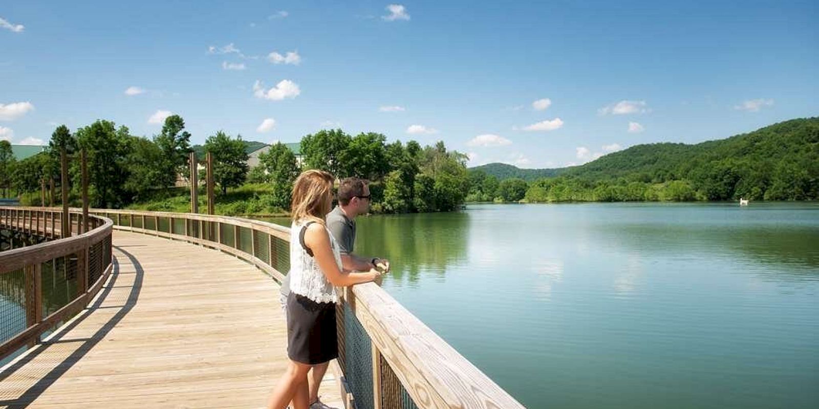 A couple stands on a lakeside boardwalk, enjoying a scenic view of calm water and green hills under a clear blue sky.