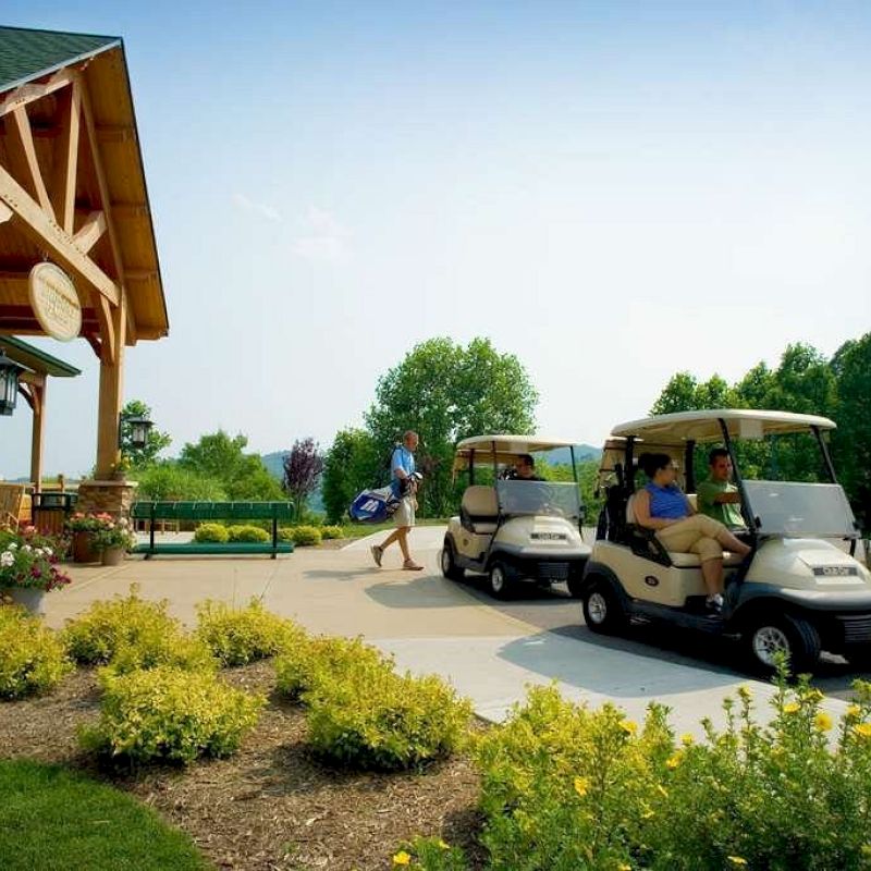 People driving golf carts near a building with green roof, surrounded by yellow flowers and trees, under a clear blue sky.