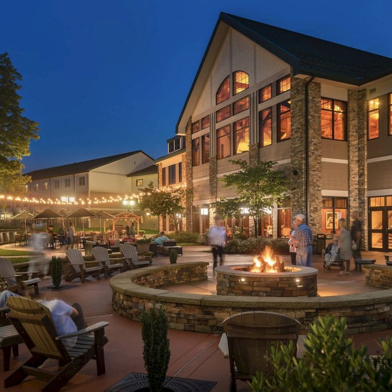 A nighttime scene of a cozy outdoor patio with a fire pit, surrounded by chairs and a lit building in the background.