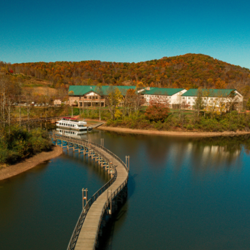 A scenic view of a lake with a curving boardwalk leading to a building, surrounded by autumnal trees and hills under a clear blue sky.