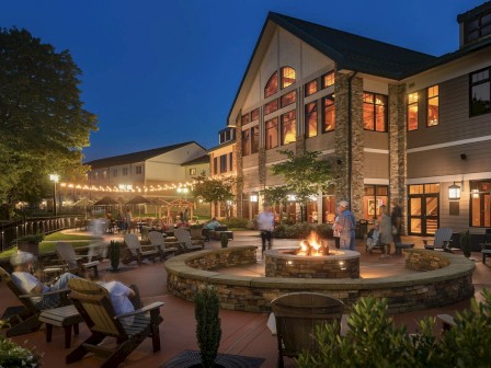 An outdoor space with a fire pit, surrounded by chairs and a building with warm lights, creating a cozy evening atmosphere.