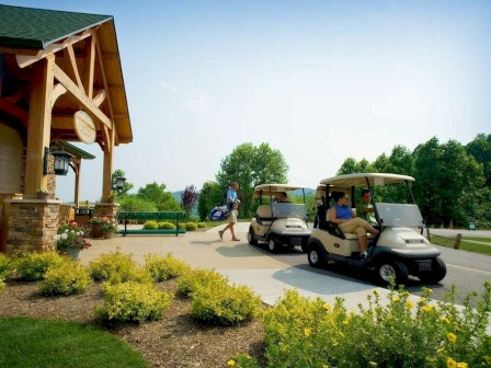 A golf course scene with two people in golf carts, another carrying clubs, near a wooden clubhouse surrounded by greenery and flowers.