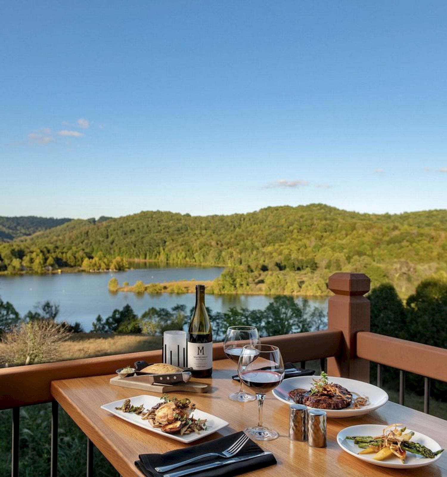A table with dishes, wine, and glasses overlooks a scenic lake and lush green hills under a clear blue sky.