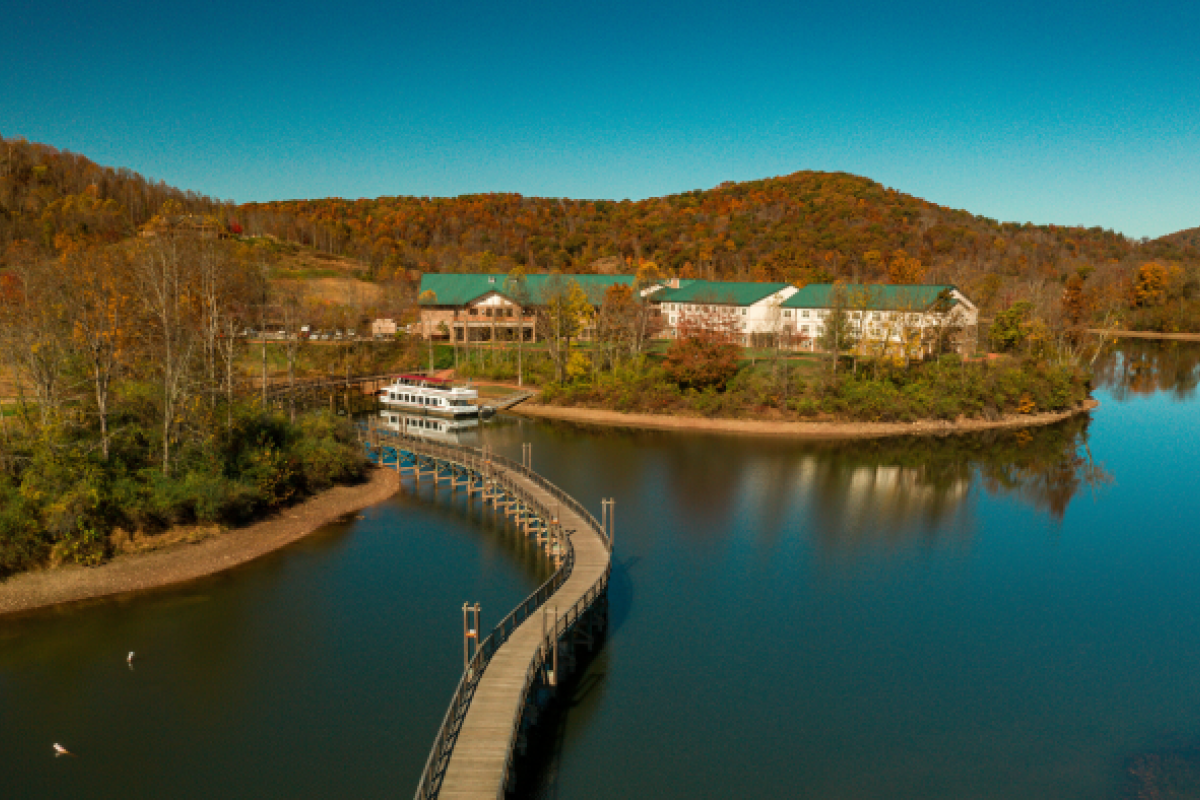 A serene landscape with a lake, a winding boardwalk, a boat, buildings, trees in autumn colors, and hills in the background.