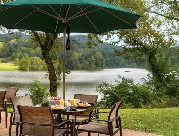 Outdoor dining setup with table, chairs, and umbrella near a lake and trees; breakfast items on the table and scenic view in the background.