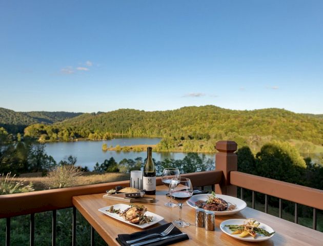 A table with meals, wine, and glasses overlooks a scenic lake and forested hills under a clear sky from a balcony.
