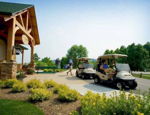 A person with a golf bag approaches as two golf carts wait outside a building surrounded by greenery and yellow flowers.