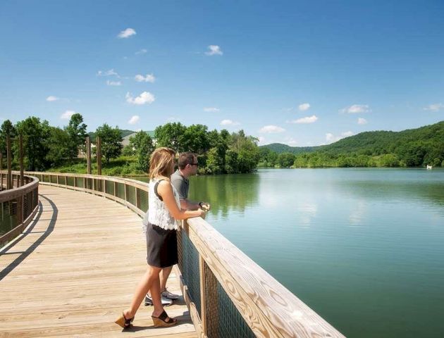 A couple stands on a wooden boardwalk, overlooking a calm lake surrounded by lush green hills and clear blue skies.