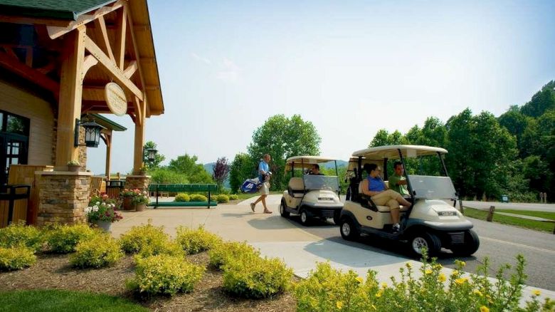 Golfers with bags and people in golf carts are outside a clubhouse, surrounded by greenery on a sunny day.