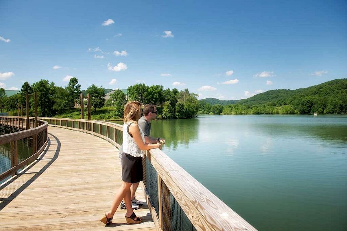 A couple is enjoying a scenic view from a wooden boardwalk over a calm lake, surrounded by lush green hills under a clear blue sky.