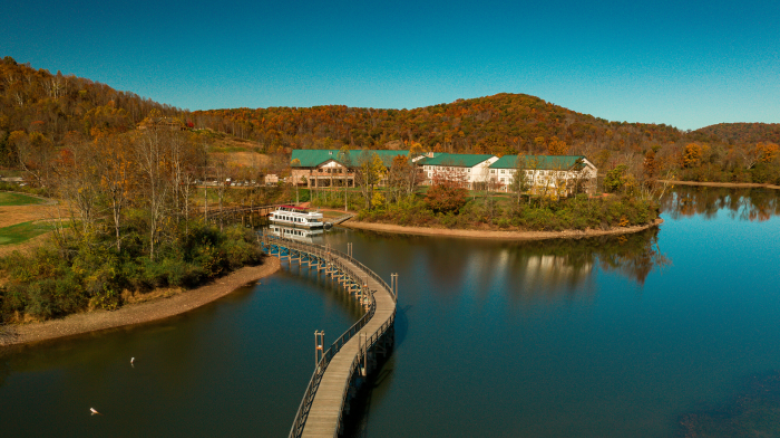 A scenic view shows a curved boardwalk over a calm lake, leading to a building complex surrounded by colorful autumn trees and hills.