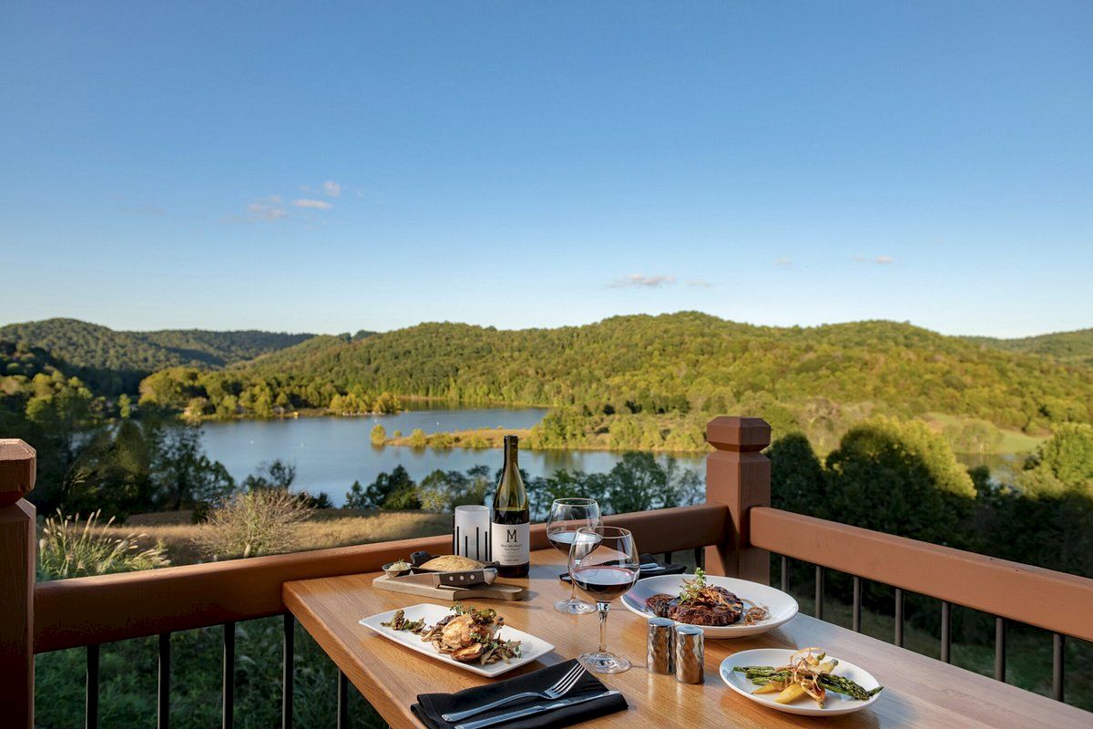 A scenic view of a lake and forest from a deck, set with plates of food, wine, glasses, and condiments on a wooden table.